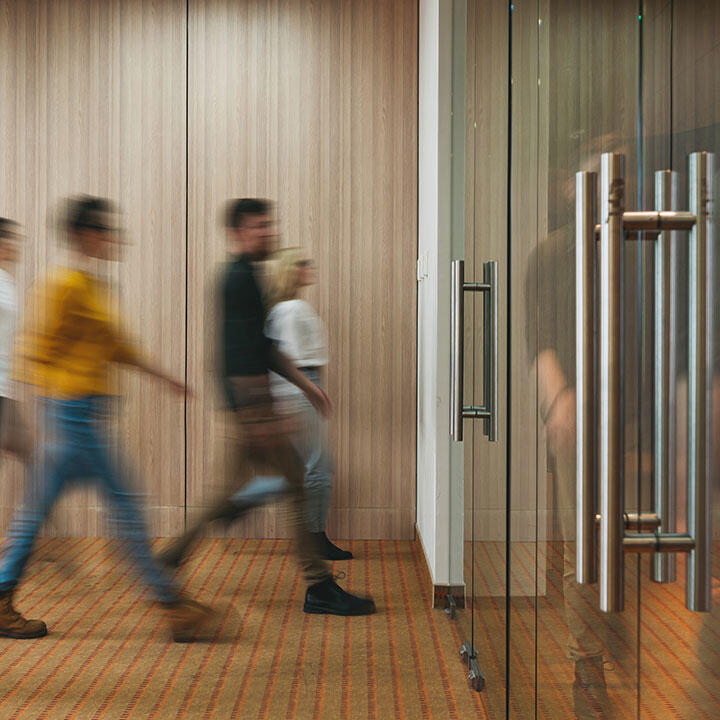 WORKARMA Blurred image of four busy people walking left-to-right to enter large glass doors.